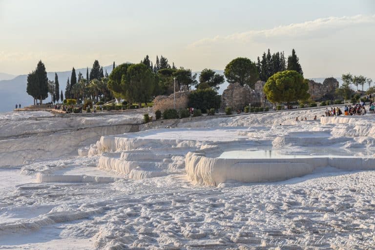 Piscinas de travertino blancas llenas de agua con una zona arbolada con ruinas al fondo y muchos turistas observando las vistas