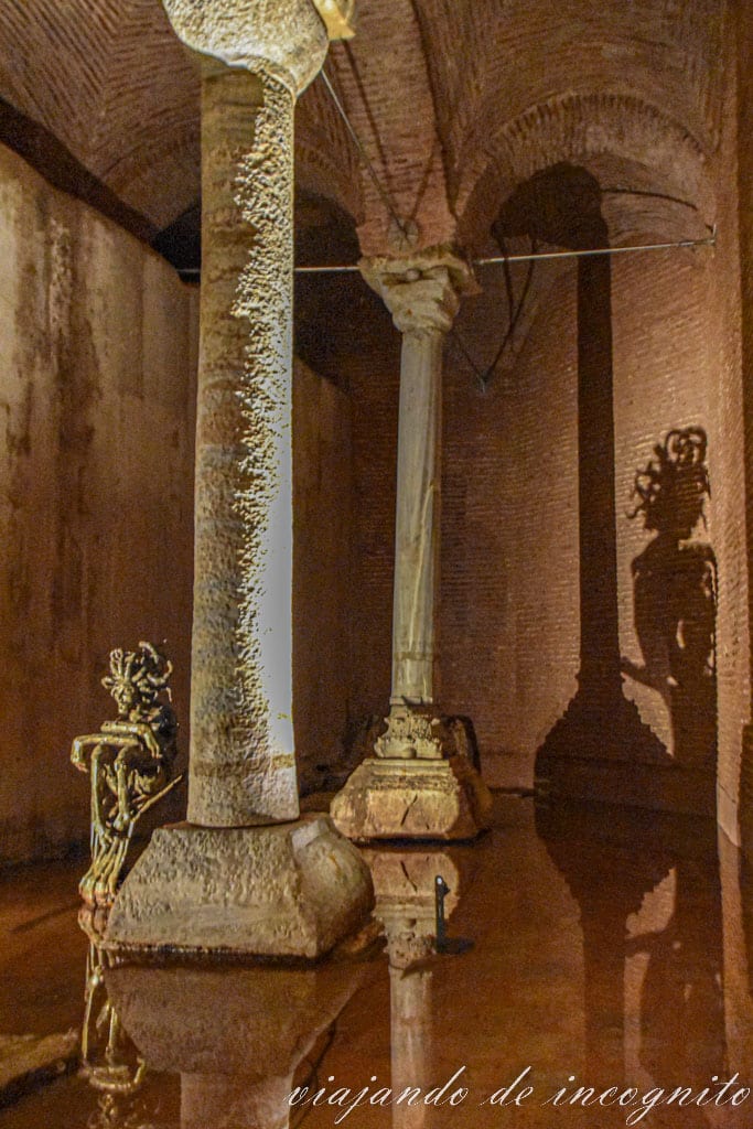 Dos columnas de la basílica cisterna junto a una escultura de un hombre sentado. Se ve el reflejo en el agua y la sombra en la pared