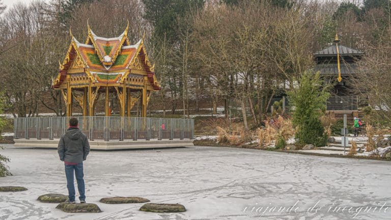 Persona de espaldas sobre una roca en un estanque congelado en invierno observando el templo tailandés con una estatua de Buddha en su interior y la pagoda nepalí junto a este en el Westpark de Múnich