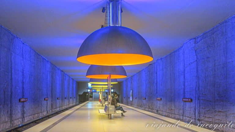 Parada de metro Westfriedhof en Múnich bañada de una luz azul y decorada con lámparas de gran tamaño del diseñador Ingo Maurer en colores naranja y amarillo