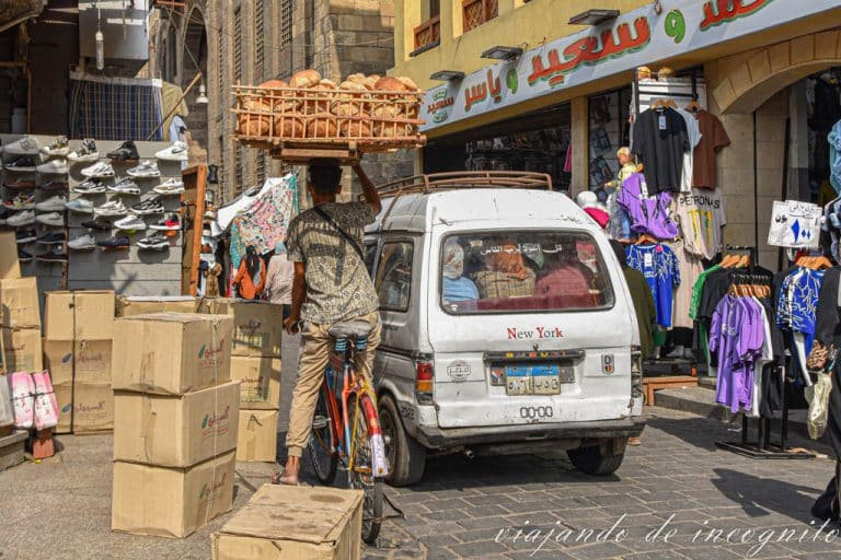 Vendedor en bicicleta transporta una bandeja llena de pan de pita sobre su cabeza mientras intenta pasar entre una furgoneta blanca y un montón de cajas