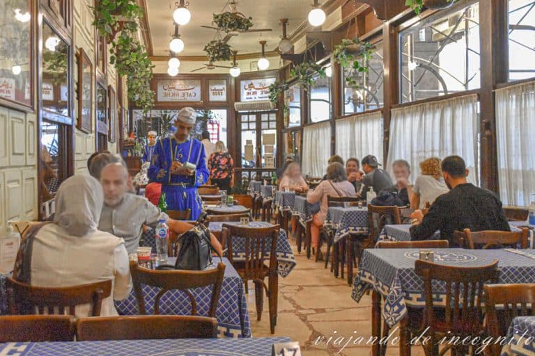 Interior del café Riche con mesas con manteles azules, clientes sentados en ellas y un camarero con ropa tradicional tomando la nota en una de ellas