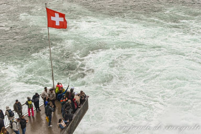 Bastante gente está en uno de los miradores de las cataratas del Rin del castillo de Laufen donde ondea una bandera suiza.