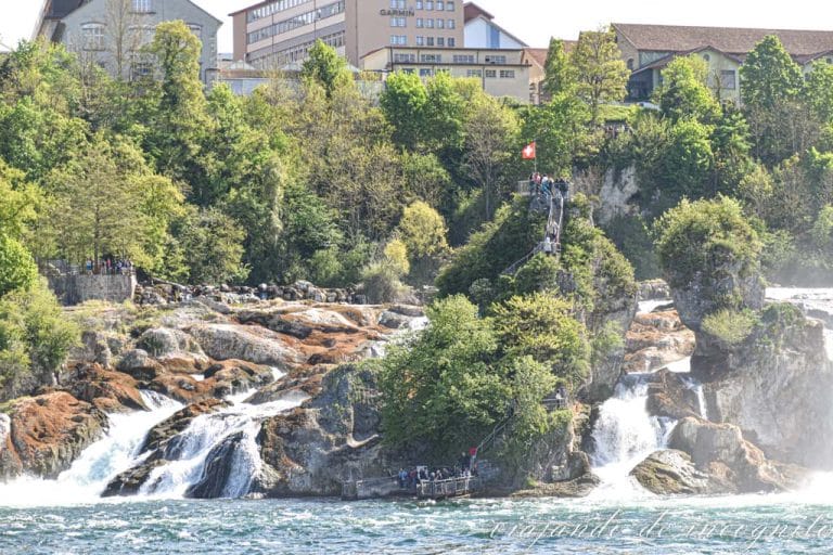 Gente subiendo por la escalera metálica del peñón que está en el medio de las cataratas del Rin y donde ondea una bandera suiza