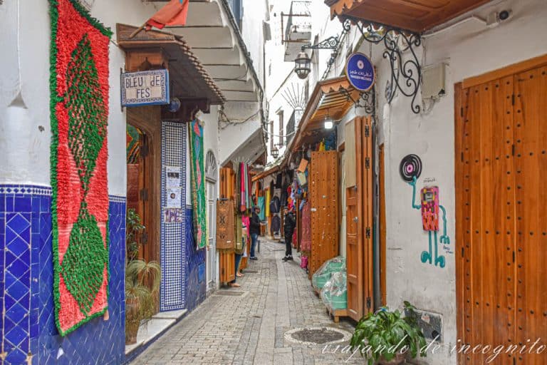 Calle de la medina de Tánger llena de tiendas y cuyas paredes están decoradas con una colorida alfombra y un grafiti.