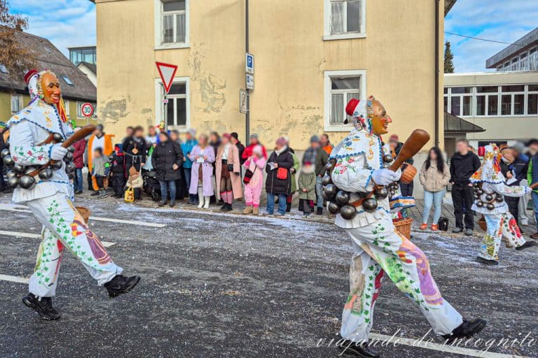 Tres personas con máscaras tradicionales y vestidas con trajes blancos pintados con grandes cascabeles desfilan por la calle principal de St. Georgen.
