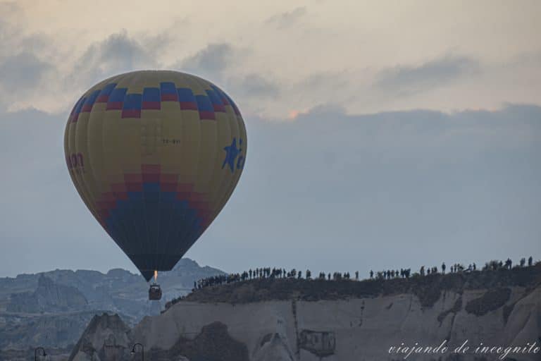 Globos amarillo con cuadros azules, rojos y naranjas volando al amanecer frente a una fila de turistas en uno de los miradores cerca de Göreme