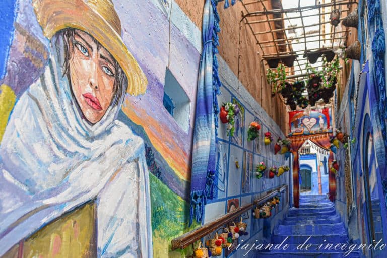 Una calle de la Medina de Chefchaouen decorada con un mural de una mujer y macetas de flores.