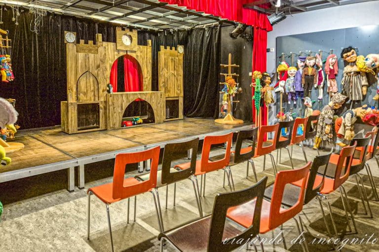 Sala del museo de las marionetas de Pilsen donde hay numerosas marionetas y un escenario para que el visitante las manipule. Frente al escenario hay dos filas de sillas vacías.
