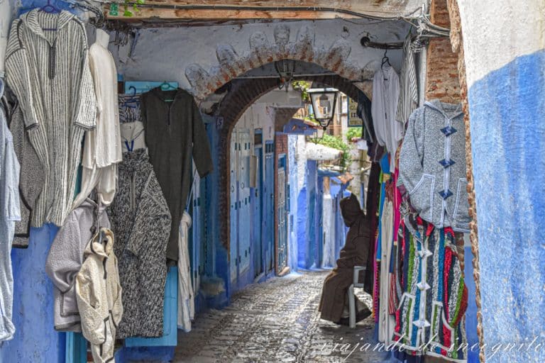 Un hombre con ropas tradicionales marroquíes está sentado bajo un arco frente a la entrada de una tienda.