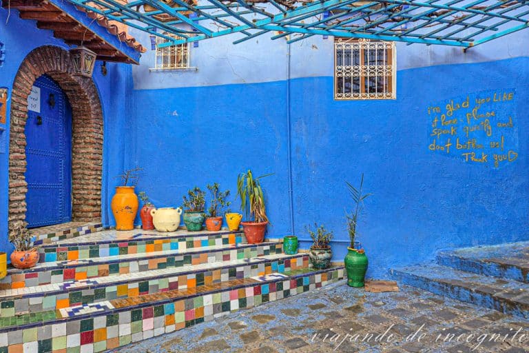 Puerta de una casa naranja con cuatro escalones frente a ella decorados con azulejos en tonos anaranjados. En la pared azul se ve escrito en inglés y en amarillo: nos alegra que te guste, pero habla bajo y no nos molestes. Gracias.