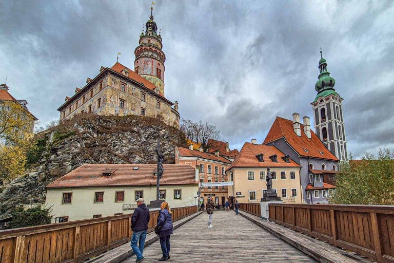 Puente de madera de Cesky Krumlov con el castillo y la iglesia de San Jost al fondo. En el puente hay una pareja mirando el paisaje.