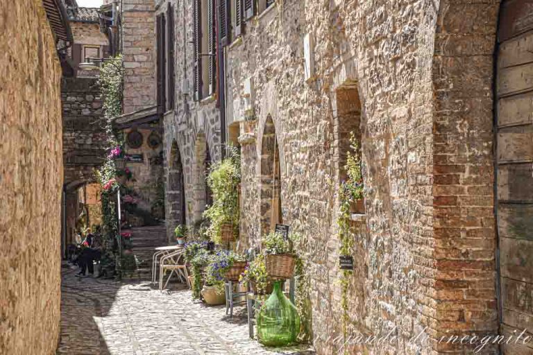 Callejón de Spello decorado con flores. Al fondo se ve un arco y a dos personas pasando por él.