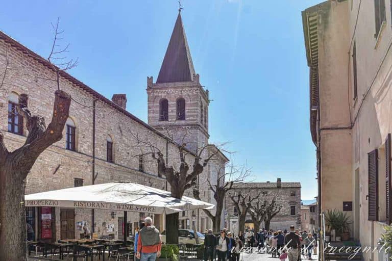 Gente paseando un día de calor por la calle donde al fondo se ve la Colegiata de Nuestra Señora la Mayor de Spello.