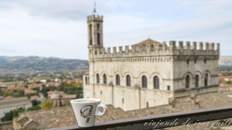 Taza de café donde pone tre ceri apoyada en una barra y de fondo Gubbio y el palacio de los Cónsules
