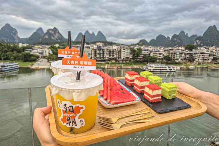 Una mano sujeta una bandeja de bambú con dos zumos, tres trozos de sandía y 6 pedazos de bizcocho. De fondo se ven las montañas de Yangshuo.