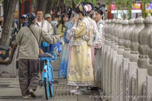 Dos mujeres vestidas en trajes tradicionales chinos hablan por el móvil mientras que una señora mayor pasa junto a ellas empujando una bicicleta.