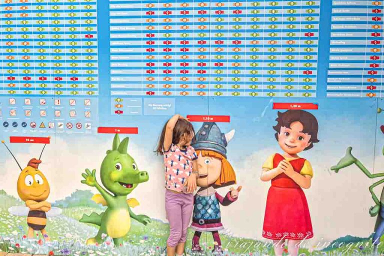 Una niña se mide en un cartel de Plopsaland donde aparecen los personajes del parque y todas las atracciones con sus restricciones de altura.